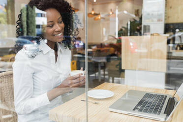 Smiling businesswoman with coffee cup looking at laptop while sitting in cafe - JRVF01570