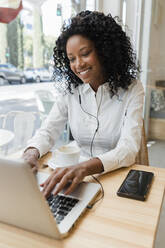 Young businesswoman with in-ear headphones using laptop at coffee shop - JRVF01579