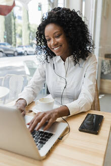 Junge Geschäftsfrau mit In-Ear-Kopfhörern und Laptop in einem Café - JRVF01579