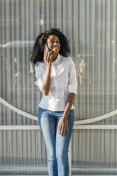 Lächelnde Geschäftsfrau, die vor einer Glaswand steht und mit einem Mobiltelefon spricht - JRVF01592