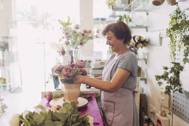 Fröhliche reife Blumenhändlerin arrangiert Blumen, während sie an einem Tisch im Geschäft steht - LJF02292