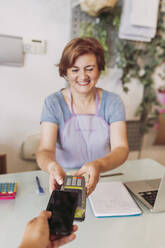 Eine lächelnde Floristin hält ein Kreditkartenlesegerät in der Hand, während ein Kunde mit seinem Smartphone an der Kasse kontaktlos bezahlt - LJF02301