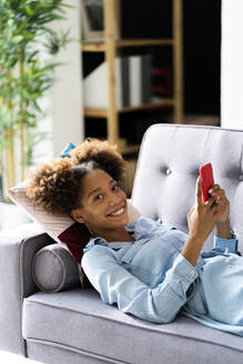 Lächelnde Frau mit Smartphone auf dem Sofa im Wohnzimmer liegend - GIOF13238