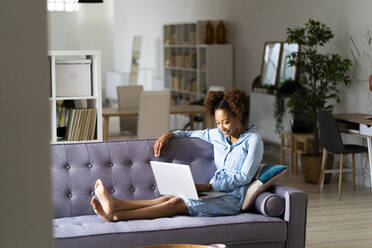 Junge Afro-Frau benutzt einen Laptop, während sie zu Hause auf dem Sofa sitzt - GIOF13243