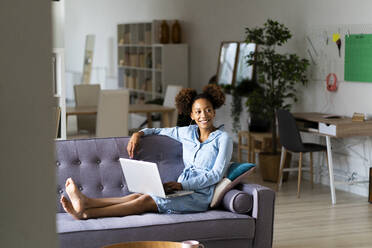 Smiling young woman sitting with laptop while looking away in living room - GIOF13244
