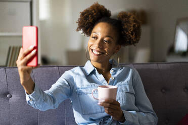 Lächelnde Frau spricht Selfie mit Kaffeetasse durch Handy zu Hause - GIOF13249