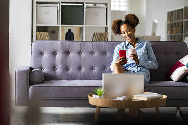 Junge Frau führt einen Videoanruf über ihr Smartphone, während sie im Wohnzimmer Kaffee trinkt - GIOF13250