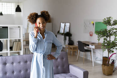 Lächelnde Frau mit Hand an der Hüfte, die im Wohnzimmer über ihr Smartphone spricht - GIOF13263