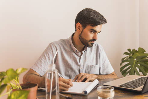 Man looking at laptop while writing in note pad - MGRF00350