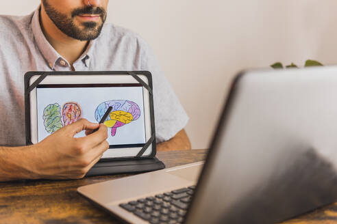 Männliche Fachkraft für psychische Gesundheit zeigt ein Gehirndiagramm auf einem digitalen Tablet während einer Online-Therapie - MGRF00369