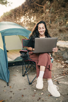 Lächelnde junge Frau, die einen Laptop benutzt und auf einem Stuhl im Wald sitzt - GRCF00850