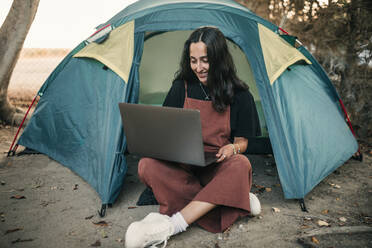 Smiling female freelancer working on laptop while sitting in tent during weekend - GRCF00852