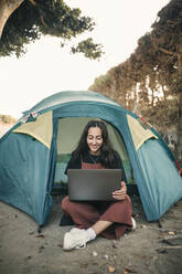 Smiling young woman working on laptop while sitting in tent during weekend - GRCF00853