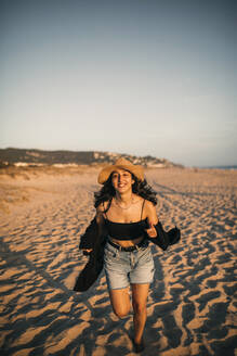 Fröhliche Frau mit Hut läuft am Strand bei Sonnenuntergang über den Sand - GRCF00856