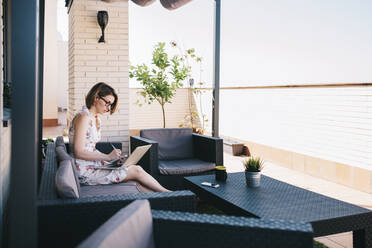 Mittlere erwachsene Geschäftsfrau mit Laptop, die bei der Arbeit auf der Terrasse in einen Notizblock schreibt - MRRF01318