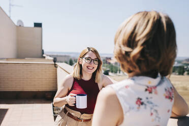 Fröhliche Geschäftsfrau, die eine Kaffeetasse hält, während sie mit einem Kollegen auf dem Dach diskutiert - MRRF01324