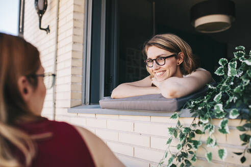 Smiling female colleague looking at businesswoman while leaning on window - MRRF01332