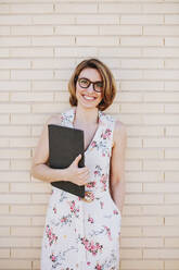 Lächelnde Geschäftsfrau mit Brille und digitalem Tablet vor einer Backsteinmauer stehend - MRRF01334