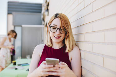 Smiling businesswoman using mobile phone while leaning on wall at rooftop - MRRF01340