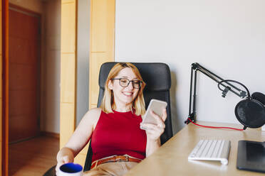 Lächelnde Geschäftsfrau, die ein Mobiltelefon benutzt, während sie auf einem Drehstuhl im Home Office sitzt - MRRF01343