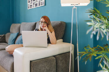 Businesswoman with hand on chin using laptop while sitting in living room - MRRF01353