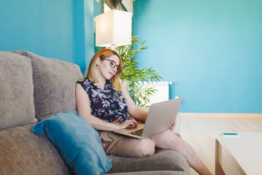 Geschäftsfrau, die einen Laptop benutzt, während sie zu Hause auf dem Sofa im Wohnzimmer sitzt - MRRF01356