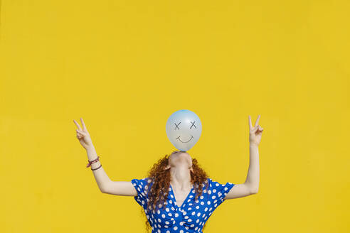 Unbeschwerte Frau balanciert Luftballon mit anthropomorphem Smiley-Gesicht vor gelber Wand - JCZF00796