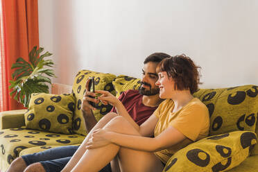 Bearded man sharing mobile phone with girlfriend while sitting on sofa in living room - MGRF00384