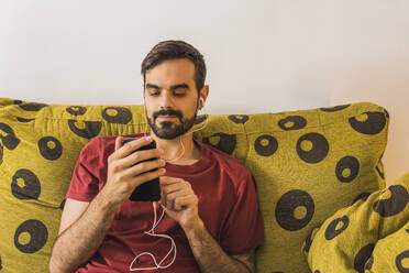 Gut aussehender Mann, der ein Mobiltelefon benutzt, während er auf dem Sofa zu Hause Musik hört - MGRF00385