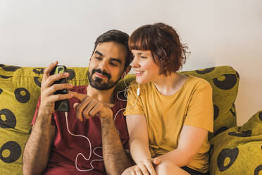 Young man sharing smart phone while listening music with girlfriend on sofa at home - MGRF00386