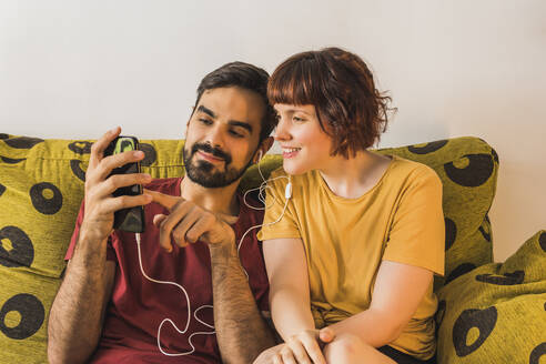 Junger Mann, der sein Smartphone mit seiner Freundin auf dem Sofa zu Hause teilt und Musik hört - MGRF00386