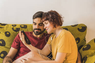 Mid adult woman sharing smart phone with boyfriend on sofa in living room - MGRF00388