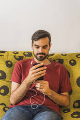 Young man using smart phone while listening music at home - MGRF00389