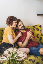 Junger Mann zeigt seiner Freundin auf dem Sofa das Mobiltelefon - MGRF00390