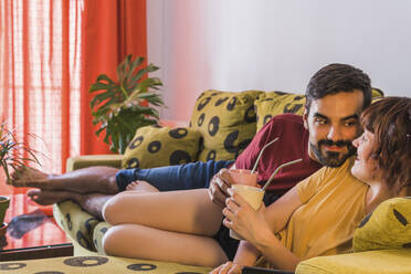 Paar mit Bambusglas, das sich gegenseitig ansieht, während es auf dem Sofa im Wohnzimmer liegt - MGRF00413