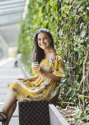 Lächelnde schöne reife Frau sitzt mit Glas vor Pflanzen - JCCMF03532