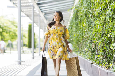 Happy mature woman with shopping bags looking away while walking on footpath - JCCMF03536