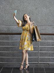 Smiling woman taking selfie with shopping bags in front of gray wall - JCCMF03539