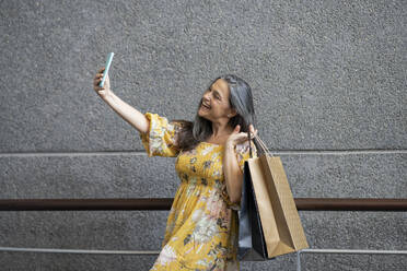 Happy woman taking selfie with shopping bags in front of gray wall - JCCMF03540