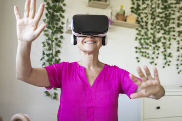 Smiling woman gesturing while using virtual reality simulator at home - JCCMF03579