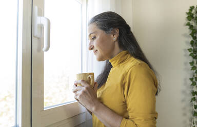 Woman looking through window while holding coffee mug at home - JCCMF03586