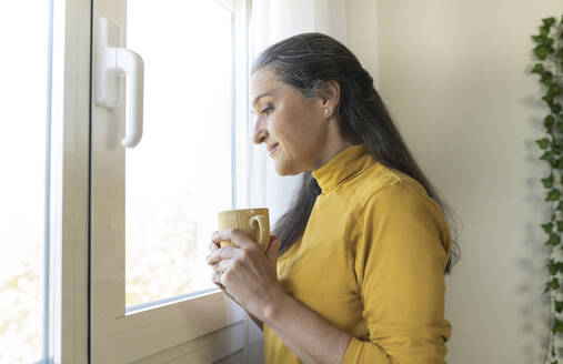 Frau schaut durch ein Fenster und hält einen Kaffeebecher in der Hand - JCCMF03586