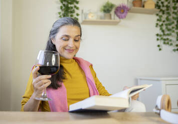 Lächelnde Frau trinkt Wein und liest ein Buch zu Hause - JCCMF03588