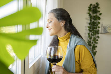 Lächelnde Frau mit Weinglas, die durch das Fenster ihres Hauses schaut - JCCMF03590