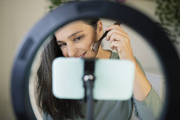 Female vlogger showing make-up tutorial seen through ring light at home - JCCMF03593