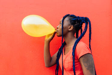 Italy, Milan, Young woman with braids blowing yellow balloon - ISF24979