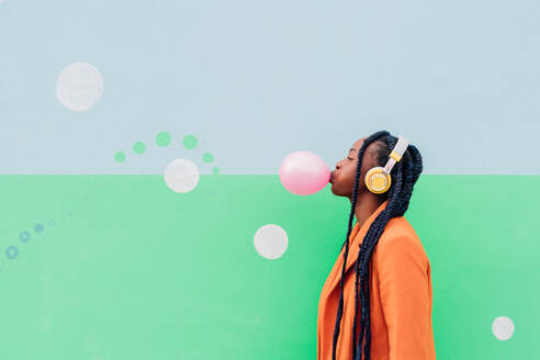 Italy, Milan, Stylish woman with headphones blowing gum against wall - ISF24992