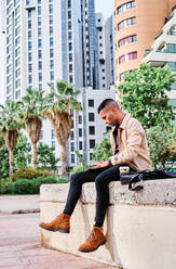 Low angle of focused young Hispanic man in stylish casual clothes working remotely on freelance project on laptop while sitting with cup of coffee near modern urban buildings - ADSF28565