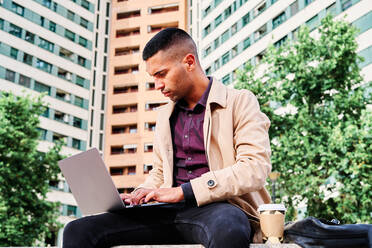 Low angle of focused young Hispanic man in stylish casual clothes working remotely on freelance project on laptop while sitting with cup of coffee near modern urban buildings - ADSF28566