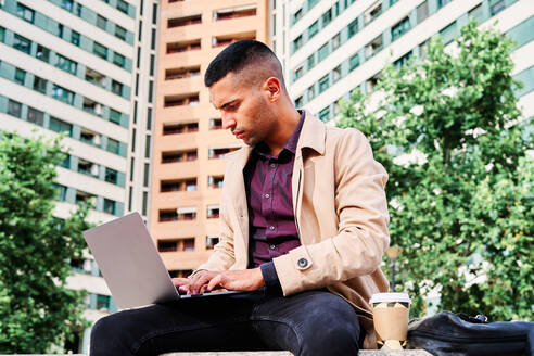 Niedriger Winkel des konzentrierten jungen hispanischen Mannes in stilvoller Freizeitkleidung, der aus der Ferne an einem freiberuflichen Projekt am Laptop arbeitet, während er mit einer Tasse Kaffee in der Nähe moderner städtischer Gebäude sitzt - ADSF28566